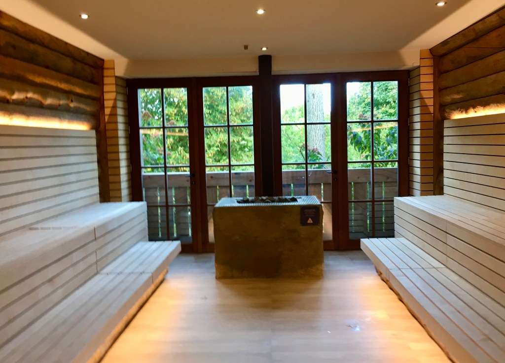 Things Helen Loves, interior of Finnish style sauna. Stove at centre, wooden benches to left and right. Floor to ceiling windows overlook woodland